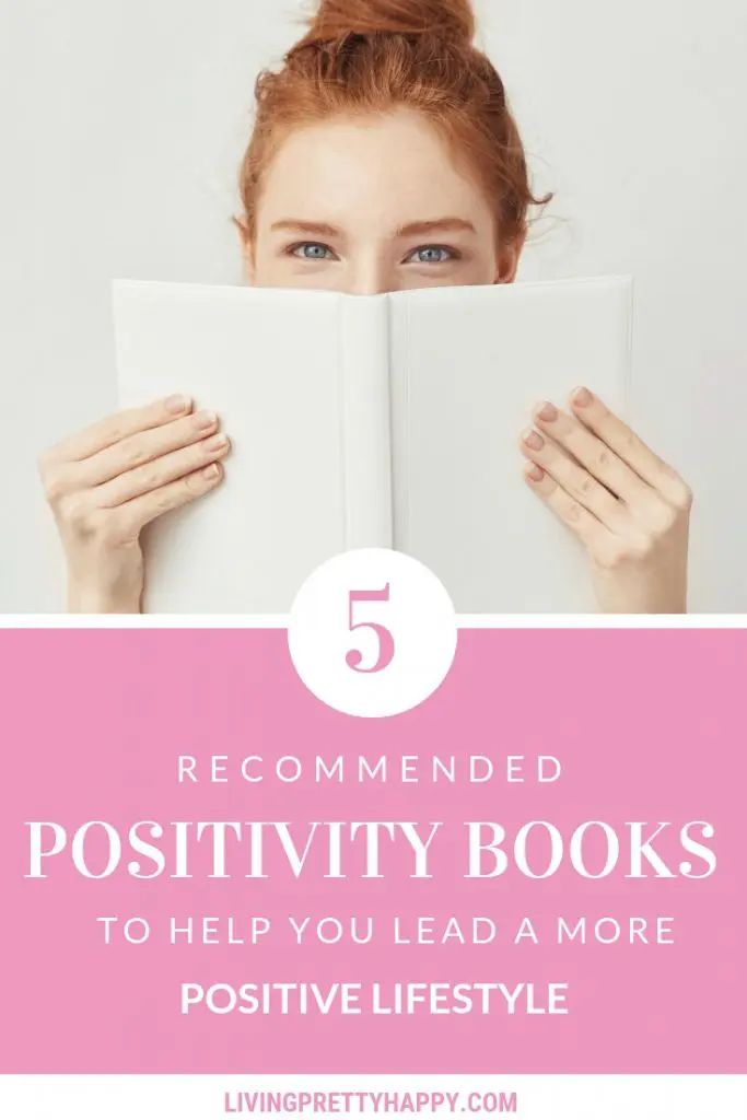 5 Recommended positivity books to help you lead a more positive lifestyle. Pinterest graphic of a ginger haired young woman holding up a blank white book over the bottom half of her face. The post title is displayed beneath the image and livingprettyhappy.com