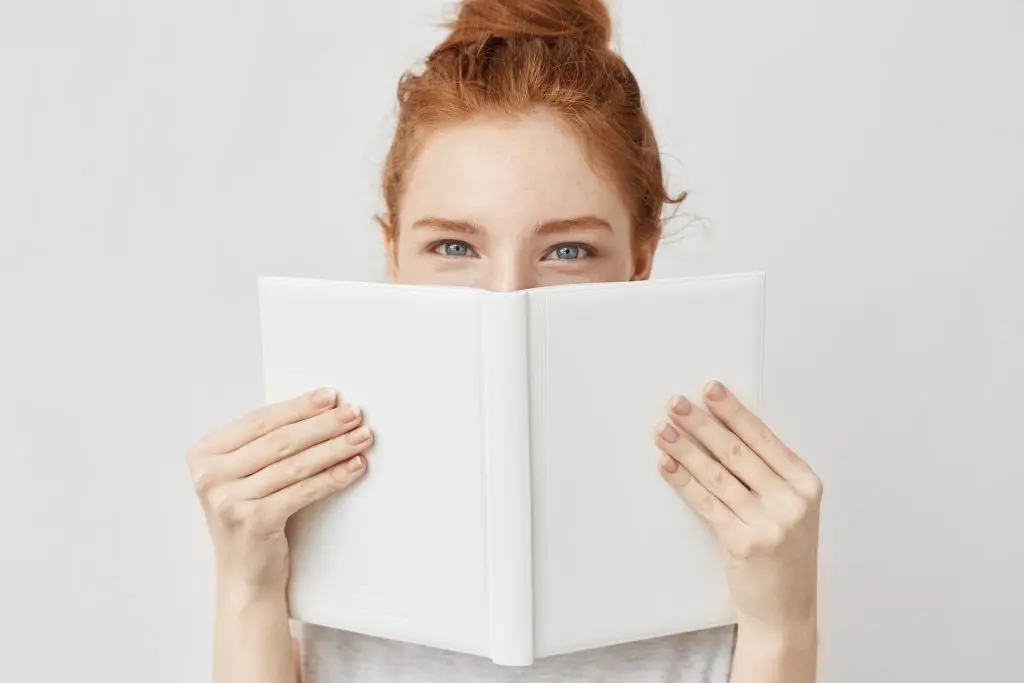 5 great books to help you lead a more positive lifestyle. Portrait of ginger girl hiding behind book looking at camera. Isolated on white background.