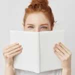 5 great books to help you lead a more positive lifestyle. Portrait of ginger girl hiding behind book looking at camera. Isolated on white background.