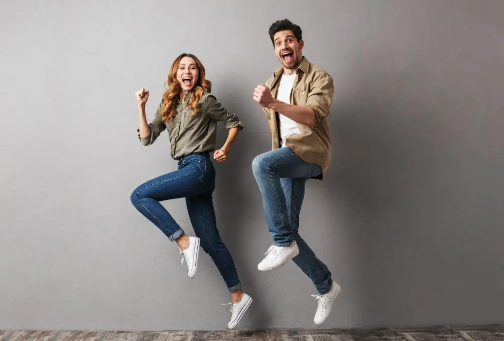 Improving well-being: The key to being happy? Image of a cheerful young couple jumping together and celebrating success isolated over gray