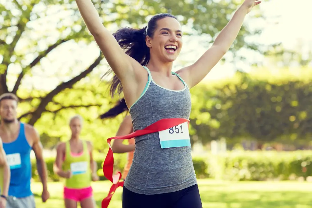How to keep your running motivation. Image of happy woman winning race and coming first to finish red ribbon over group of sportsmen running marathon with badge numbers outdoors