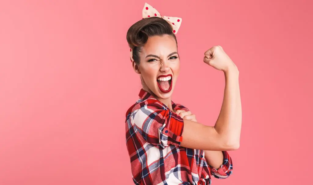 Exercise Hacks: How to be fitter without really trying. Image of young woman pulling a determine smiling face whilst showing her bicep as a display of strength on a pink background