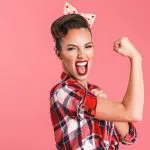 Exercise Hacks: How to be fitter without really trying. Image of young woman pulling a determine smiling face whilst showing her bicep as a display of strength on a pink background