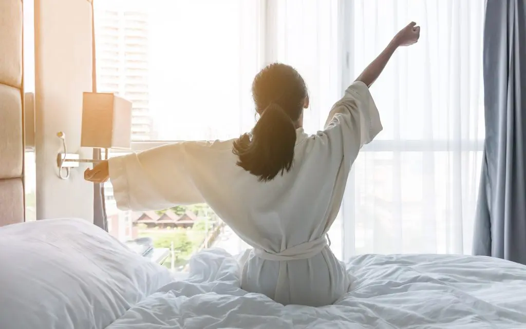 Sleep well: How to win at sleeping! (and why you should want to). How to get a good night's sleep. Image of young woman waking up, stretching out her arms, facing a floor to ceiling window with light pouring in