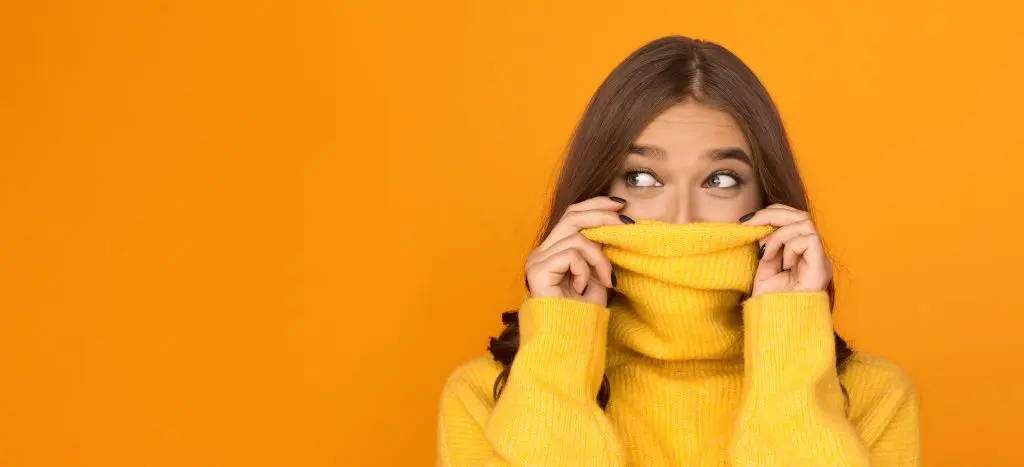 I’m new here: 10 ways to feel more confident in new situations. Image of woman hiding her face partially in her yellow jumper against an orange background