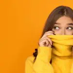 I’m new here:  10 ways to feel more confident in new situations. Image of woman hiding her face partially in her yellow jumper against an orange background