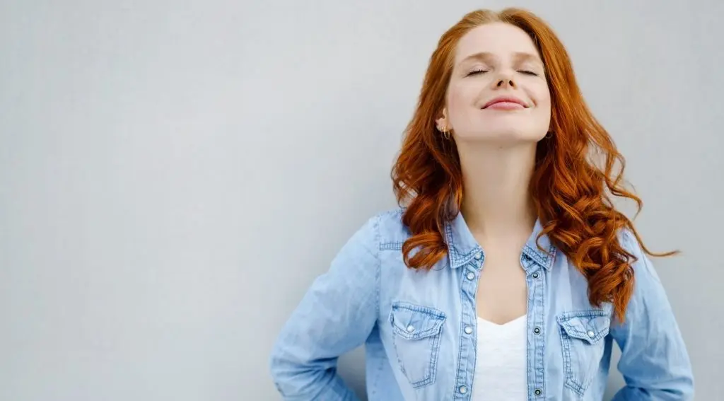 Maintaining the (inner) peace: 5 products to help calm & increase your well-being. Image of a young red-haired woman leaning against the wall, eyes closed and smiling looking at peace.