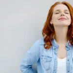 Maintaining the (inner) peace: 5 products to help calm & increase your well-being. Image of a young red-haired woman leaning against the wall, eyes closed and smiling looking at peace.