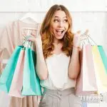 How can clothes make you happy? Plus expert tips for more successful shopping. Image of Happy young woman holding shopping bags while standing at the clothing store