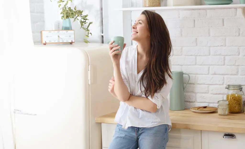 5 Ways to build self-worth. A contented woman stands happily in her kitchen drinking from a mug
