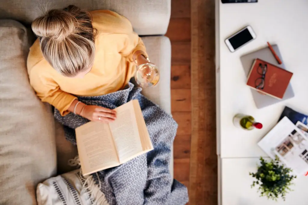 A young woman sits taking time out for herself reading a book and drinking a glass of wine
