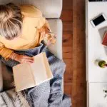 A young woman sits taking time out for herself reading a book and drinking a glass of wine