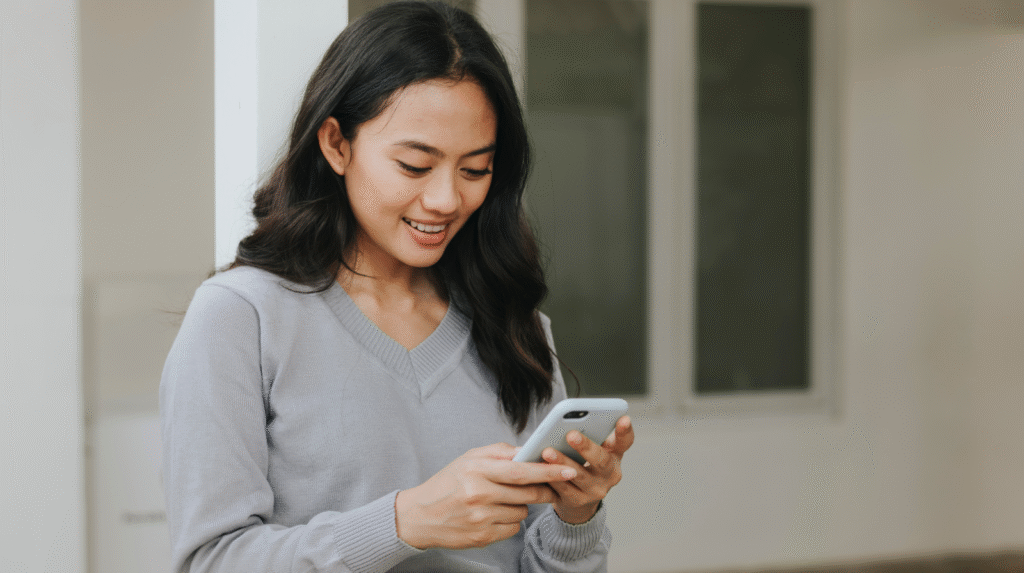 A young woman smiling while using a smartphone, dressed in a gray sweater, standing indoors with light-colored walls. Set a gentle reminder to ensure you always get your micro break benefits