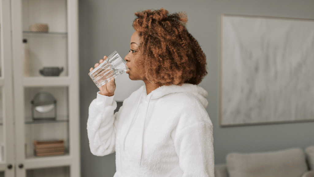 A person wearing a white hoodie is drinking water from a glass in a cozy indoor setting. Mindful hydration and nourishment is one of 10 sustainable self-care tips