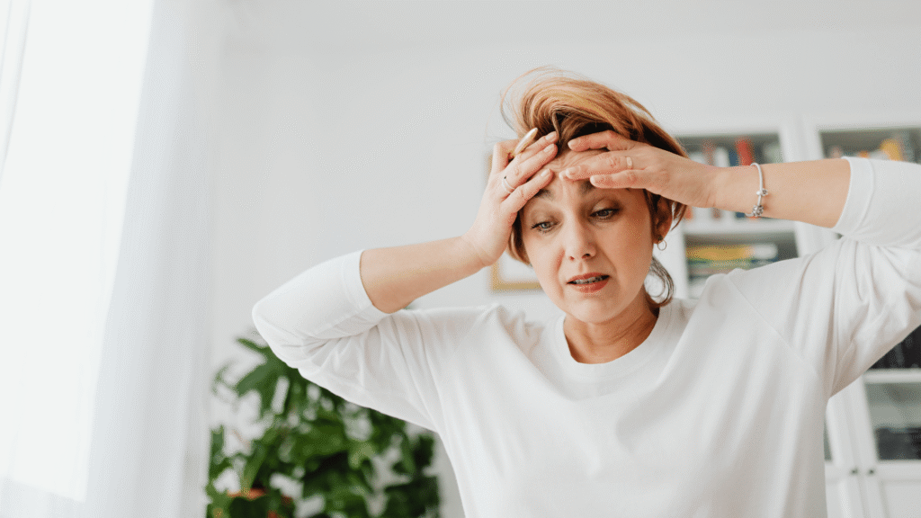 A woman in a white shirt looks stressed, holding her head with both hands, conveying feelings of overwhelm or anxiety in a bright indoor setting. Self-care is supposed to calming but if done wrong it has the opposite effect.