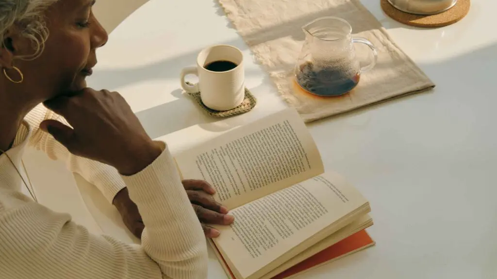 An image of a woman sitting at a table relaxing reading a book with a coffee beside her to illustrate how to calm your nervous system
