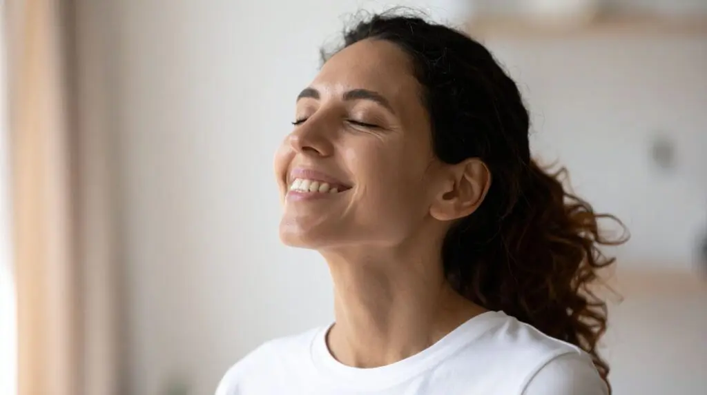 woman in a white t-shirt, hair back in a pony tail with eyes closed and smiling looking content.