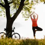 Woman in a field under a tree with a bicycle resting against it. Woman is holding a yoga pose, demonstrating the link between good mental health and a balance lifestyle