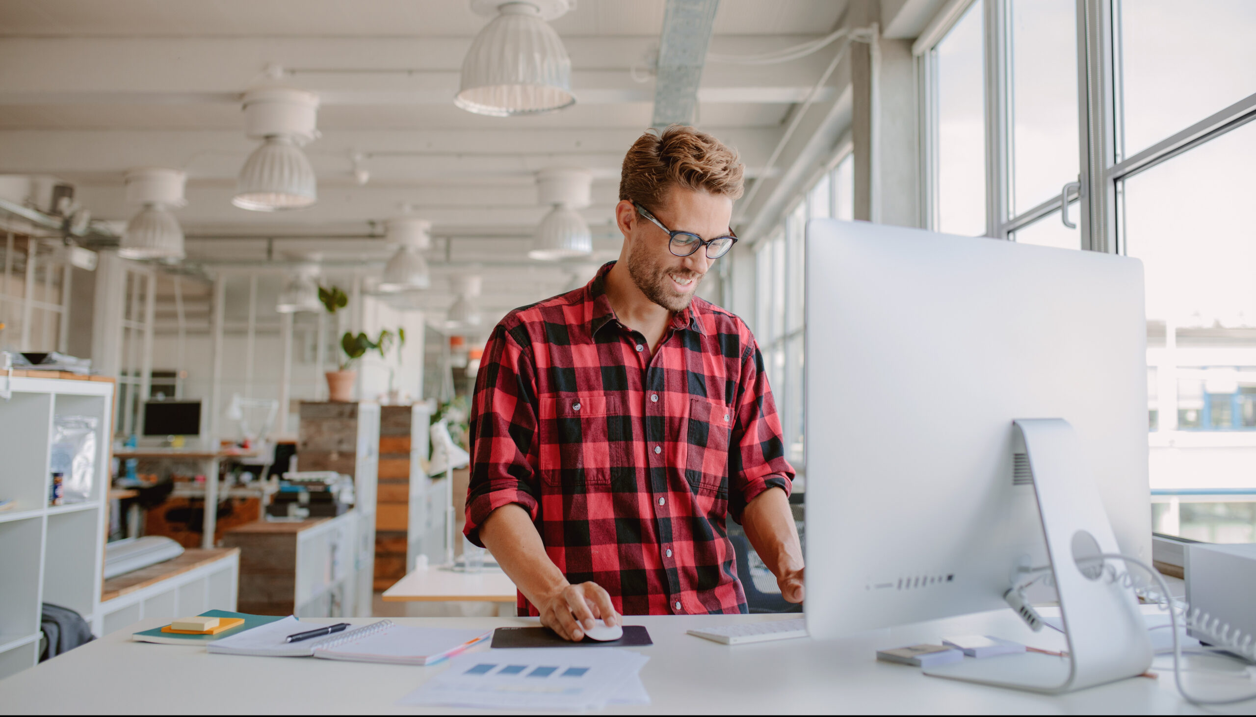 Exercise Hacks: How to be fitter without really trying. Image of Shot of happy young man standing up working on desktop computer in modern workplace.