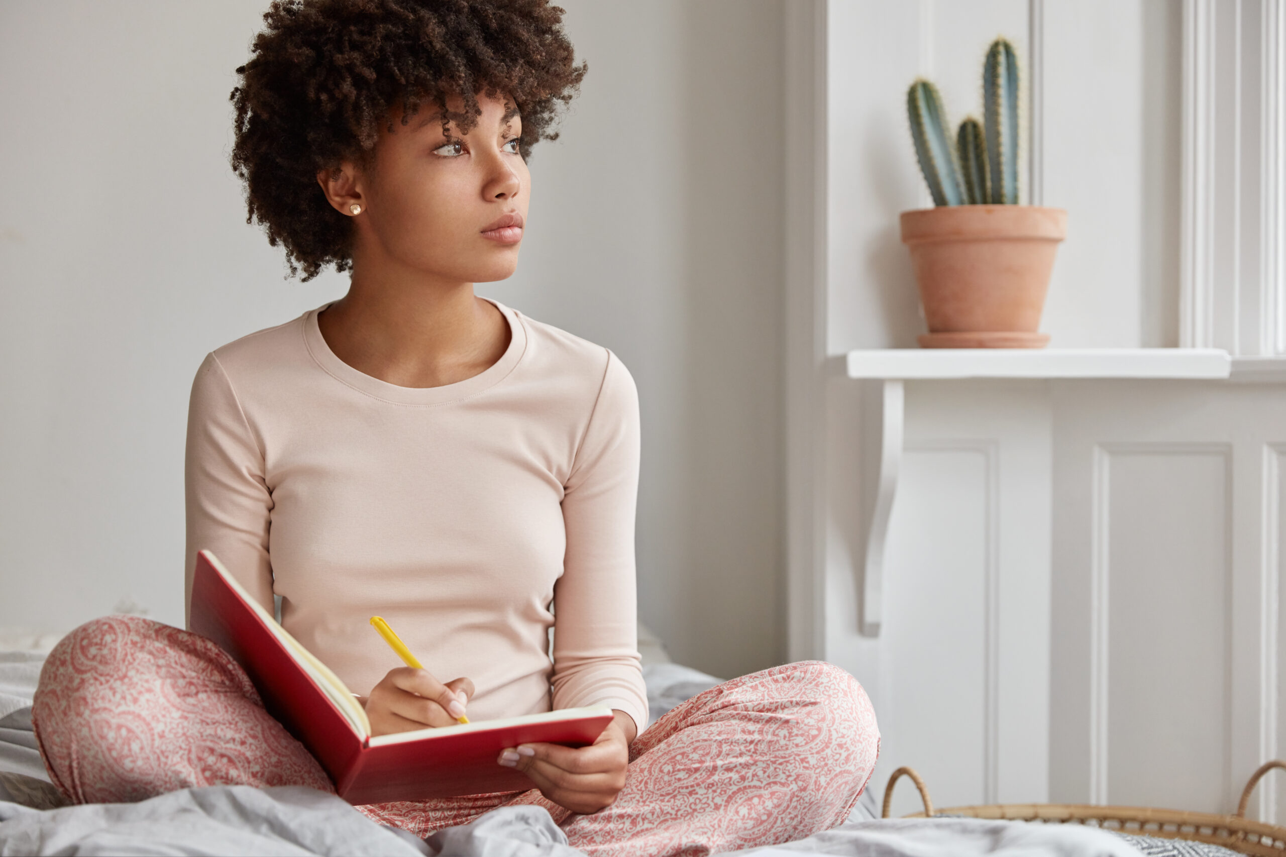 decatastrophizing - how to calm your worrying thoughts. Image of Cropped shot of dark skinned woman sits crossed legs, wears pyjamas, makes notes in diary, focused aside, blank copy space against domestic interior. Blogger creates publication for blog in notebook